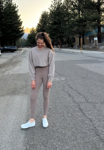 Sudadera recortada gris claro, leggings beige de cintura alta y zapatillas blancas sin cordones, situadas en un camino flanqueado por árboles durante el atardecer.