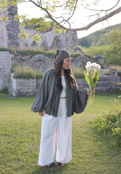 Mujer con chaqueta gris holgada y pantalones blancos de pie sobre la hierba, sosteniendo dientes de león, con ruinas de piedra y árboles al fondo.