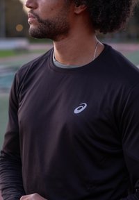 Mann mit lockigem Haar und Bart, der ein schwarzes, langärmeliges Sportshirt mit einem weißen Asics-Logo in Brusthöhe im Freien trägt.