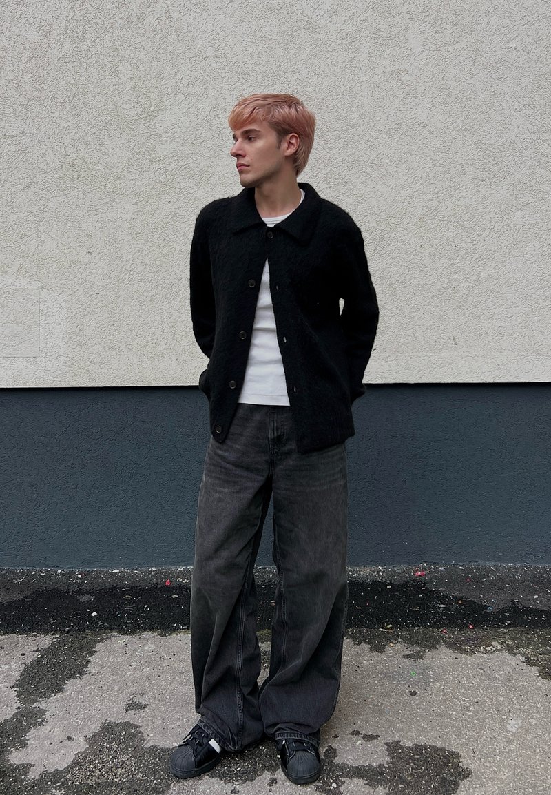 Jeune personne aux cheveux rose clair portant un cardigan noir, une chemise blanche, un jean large foncé et des baskets noires, debout contre un mur texturé.