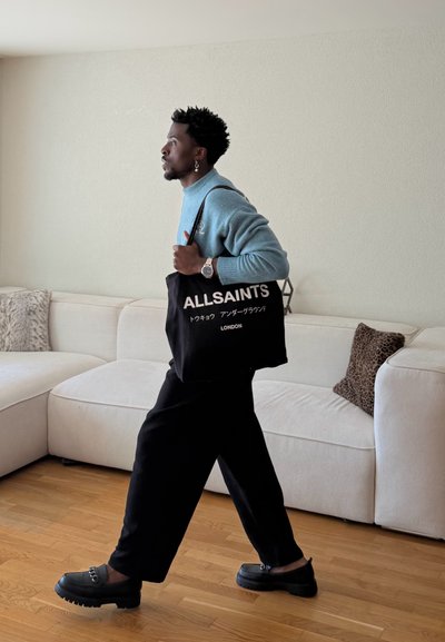 Hombre caminando en interior llevando una bolsa tote negra con el texto "ALLSAINTS LONDON", vistiendo un suéter azul, pantalones negros y zapatos negros.