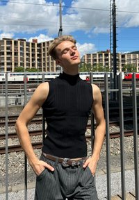 Melns, bez piedurknēm ar riboti faktūru un augstu apkakli tops; kombinēts ar pelēkiem svītrainiem biksēm un dekoratīvu brūnu jostu. Āra vide.