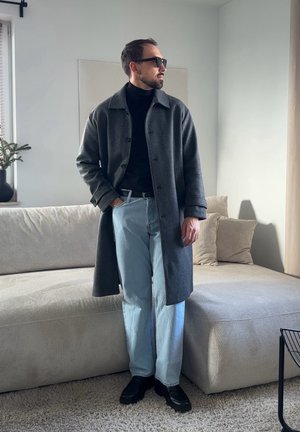 Hombre con abrigo gris oscuro, suéter de cuello alto negro, vaqueros azul claro y zapatos negros, de pie junto a un sofá en una sala de estar luminosa.