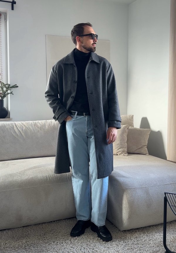 Mężczyzna w ciemnoszarym płaszczu, czarnym golfie, jasnoniebieskich dżinsach i czarnych butach, stojący obok sofy w jasnym salonie.