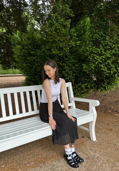 Falda negra con pliegues, top sin mangas blanco y sandalias negras con tiras. Sentada en un banco blanco en un entorno verde al aire libre.