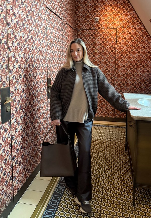 Mujer con cabello largo y rubio, vestida con una chaqueta a cuadros, suéter beige y pantalones negros, sosteniendo un bolso grande y oscuro en una habitación con patrones.