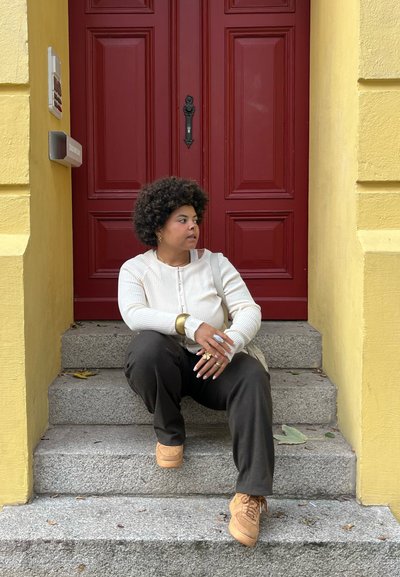 Suéter beige de canalé, pantalones gris oscuro y zapatillas color beige. Sentado en unos escalones de piedra frente a una pared amarilla y una puerta roja. Joyas notables como acento.