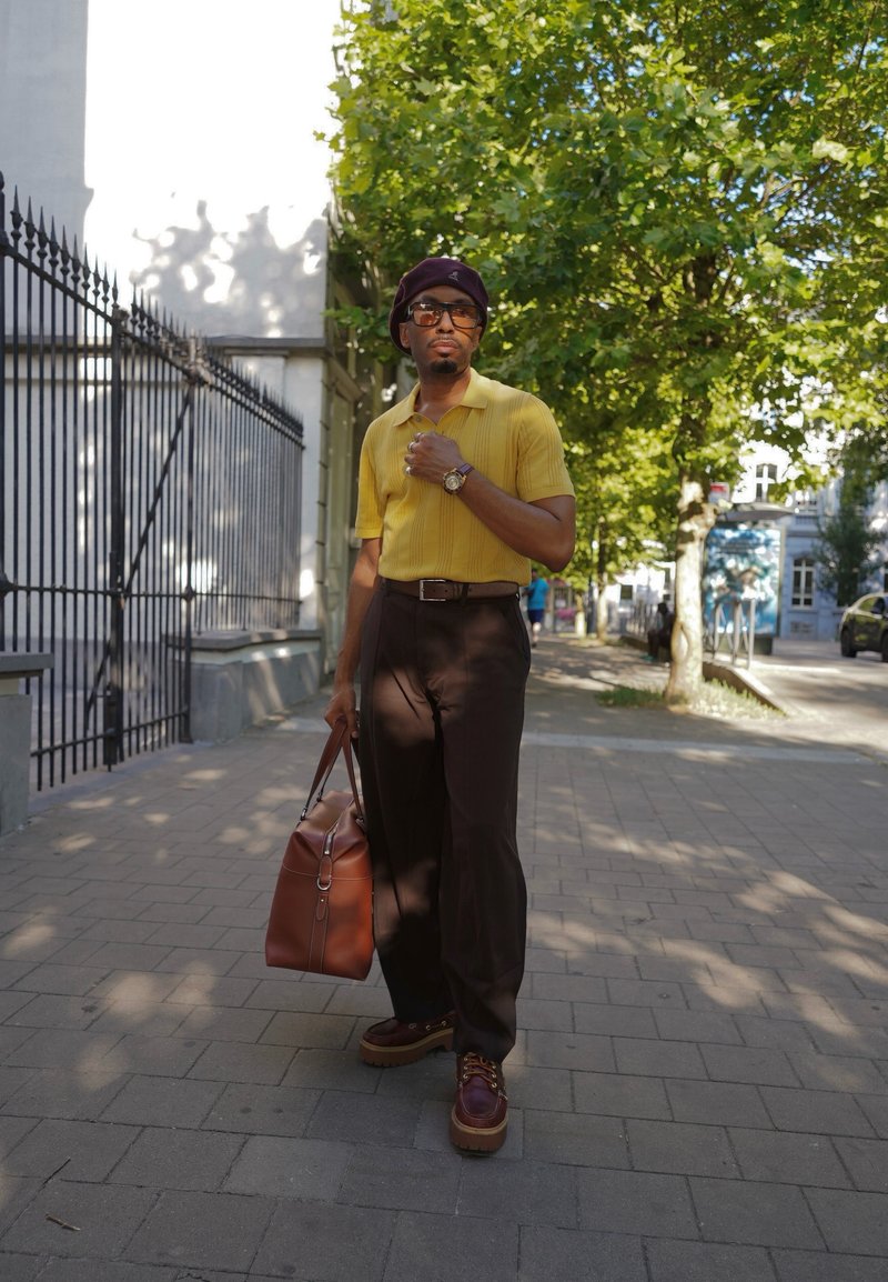 Homme portant une chemise jaune, un pantalon foncé et un béret, tenant un sac en cuir marron, debout sur un trottoir ensoleillé de la ville avec des arbres et une clôture métallique à proximité.