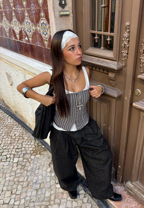 Joven mujer que lleva un chaleco a rayas y pantalones anchos negros está junto a una pared de azulejos con patrón y una puerta de madera marrón ornamentada.