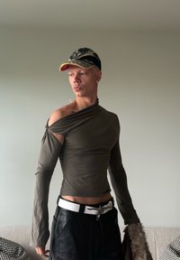 Top largo de color verde oliva, con un diseño asimétrico que presenta un hombro descubierto y una forma ajustada. Pantalones de cuero negros y una gorra.