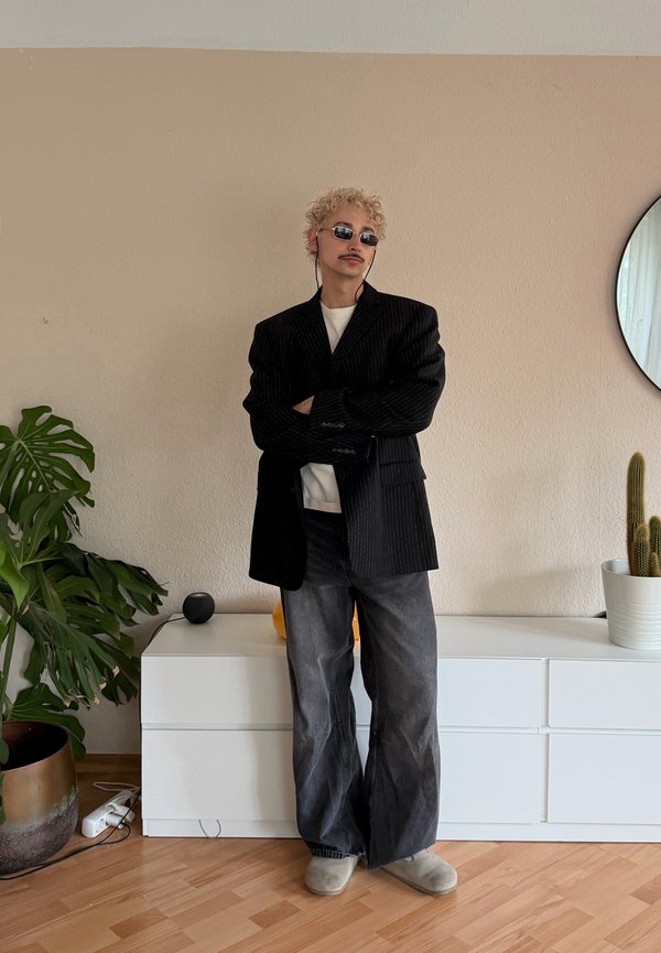 Person mit lockigem blondem Haar und Sonnenbrille steht mit verschränkten Armen, trägt einen schwarzen übergroßen Blazer und weite graue Jeans in einem minimalistischen Raum.