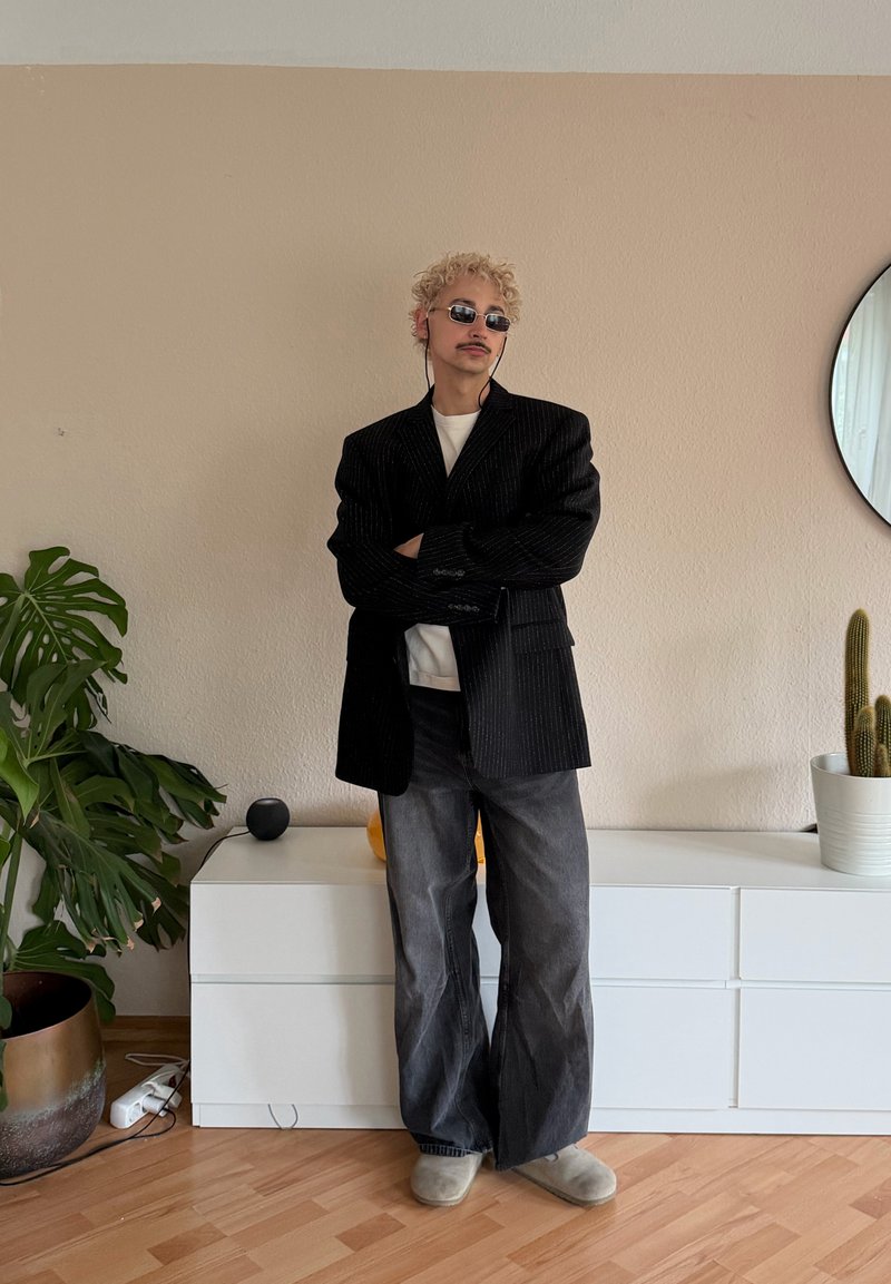 Personne aux cheveux blonds bouclés et lunettes de soleil, debout les bras croisés, portant un blazer noir oversize et un jean gris large dans une pièce minimaliste.