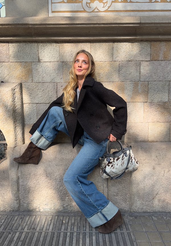 Mujer con pelo rubio largo, con chaqueta negra y vaqueros anchos remangados, sentada en un saliente de piedra, sosteniendo un bolso con estampado blanco y negro.