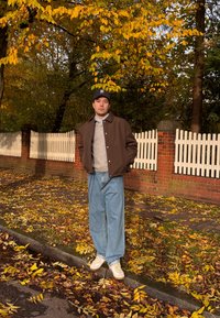 Jachetă maro peste un pulover gri, blugi albaștri largi și teniși albi. Stă pe o stradă plină de frunze, cu un gard alb de lemn în fundal.