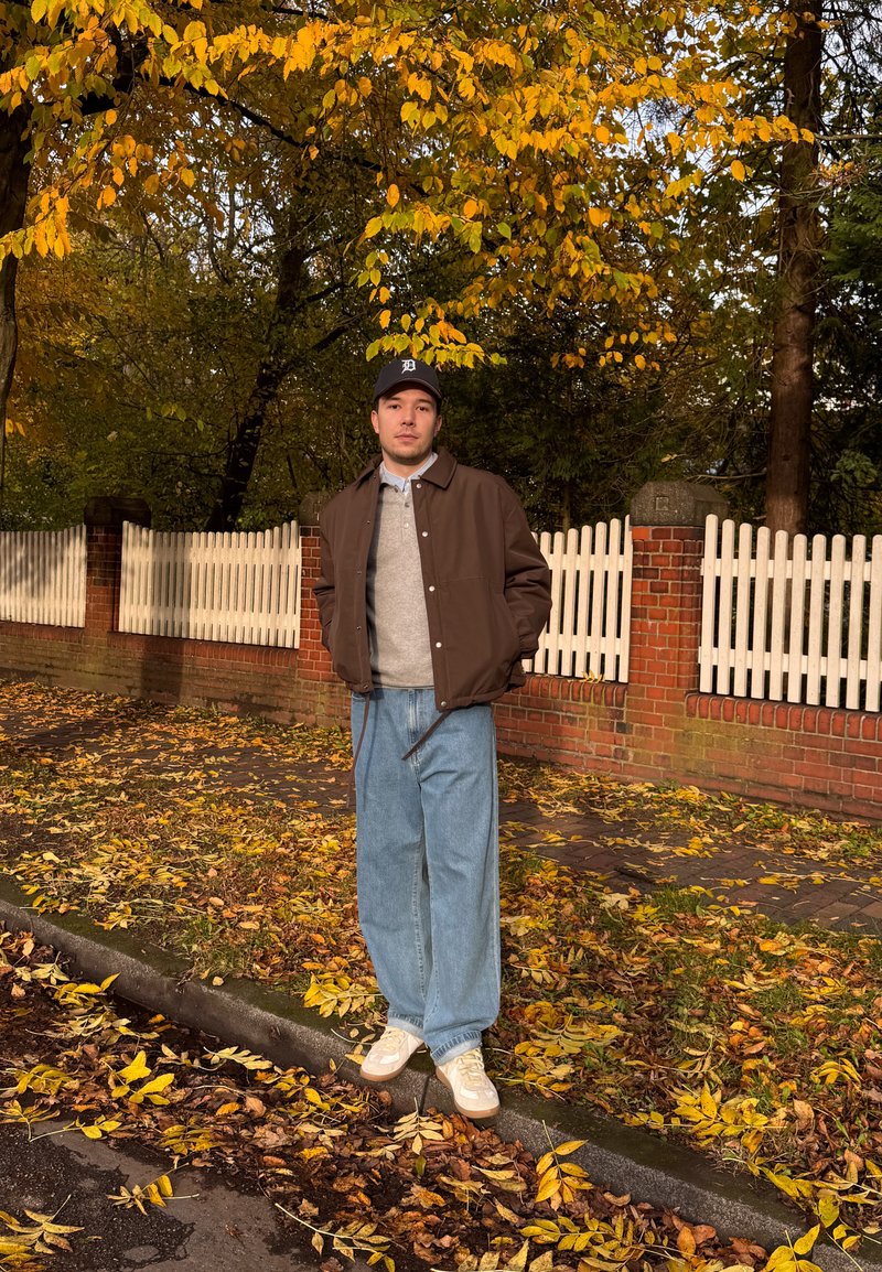 Brun jakke over en grå sweater, løse blå jeans og hvide sneakers. Står på en bladfyldt gade med en hvid stakit som baggrund.
