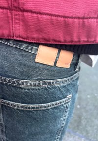 Persona che indossa jeans di denim blu con toppe in pelle color cuoio sulla cintura e una giacca rossa sopra.