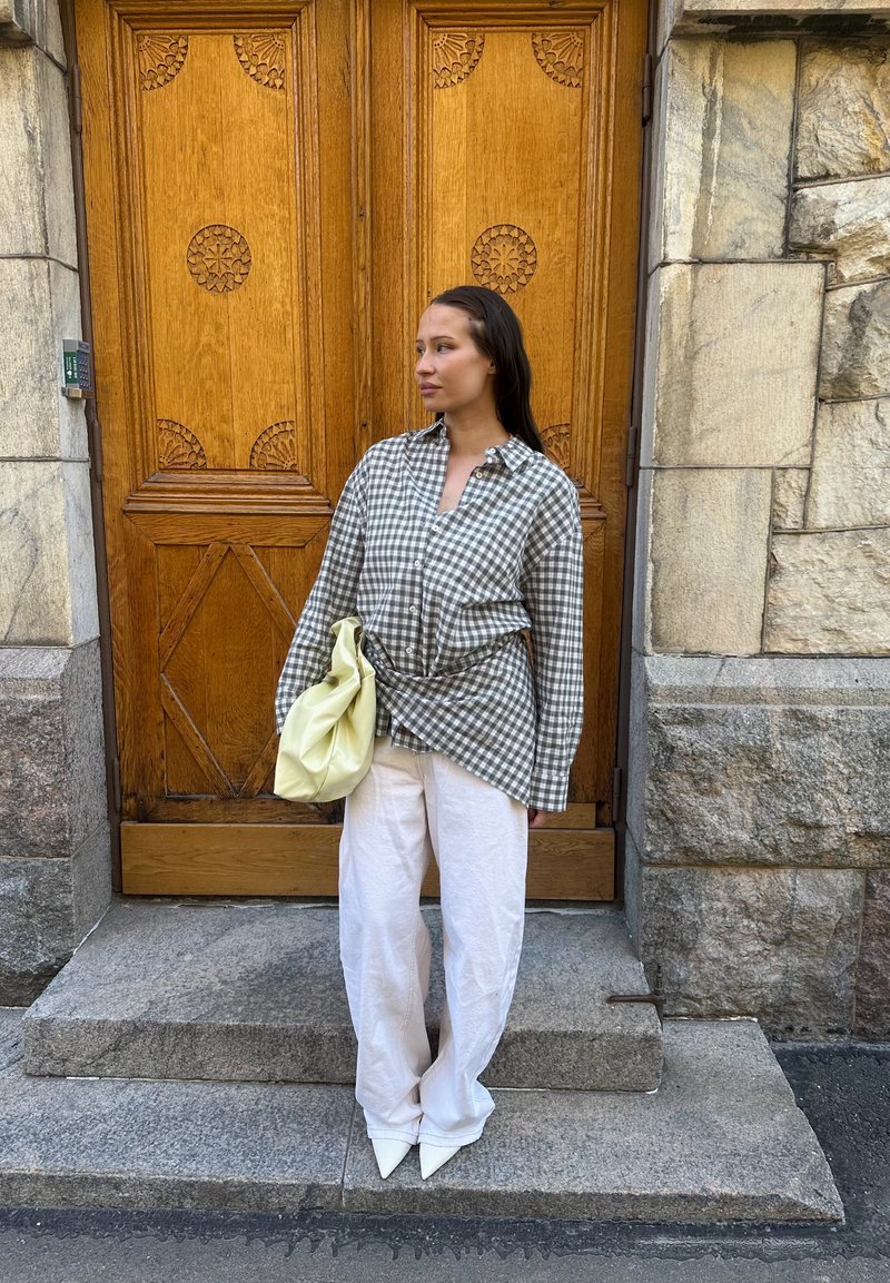 Femme aux longs cheveux foncés debout sur des marches en pierre devant une porte en bois sculptée, portant une chemise à carreaux, un pantalon blanc, des chaussures pointues et tenant un sac jaune.