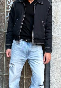 Uomo con giacca nera sopra una maglietta nera e jeans azzurri strappati, in piedi contro una parete di blocchi di vetro e una superficie grigia texturizzata.