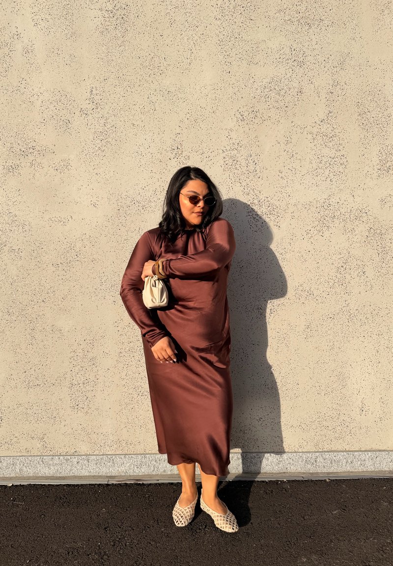 Moteris su vidutinio ilgio tamsiais plaukais, dėvi rudą satino suknelę, smėlio spalvos austus batus, saulės akinius, laiko mažą smėlio spalvos rankinę, stovinti prie tekstūruotos sienos.