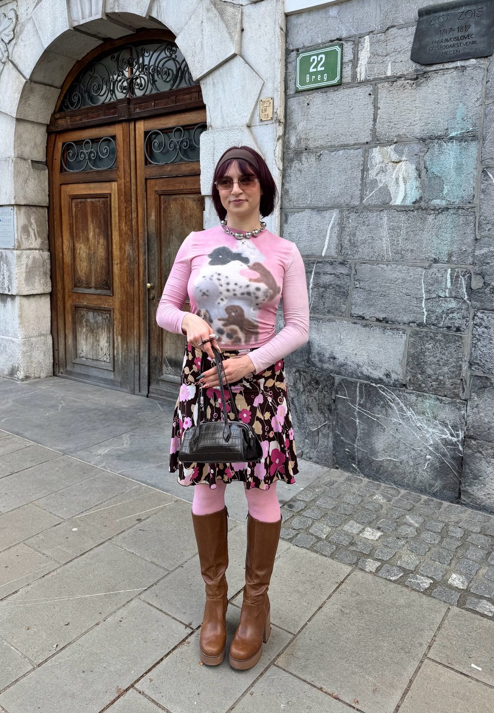 Blusa de manga longa rosa com um padrão colorido, saia preta com flores, collants rosa, botas de plataforma castanhas e uma pequena bolsa preta.