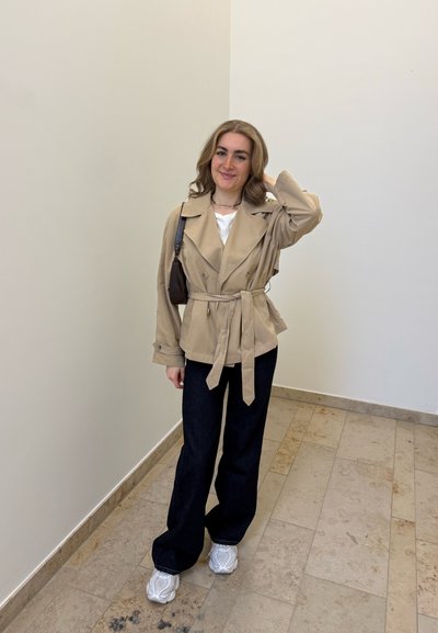 Joven mujer sonriendo, lleva chaqueta beige con cinturón, pantalones negros, zapatillas blancas, de pie en una esquina con paredes neutras y suelo de baldosas.