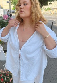 Λευκό, υπερμεγέθη πουκάμισο σε ζαρωμένο ύφασμα, φορεμένο πάνω από γυμνό τοπ. Ξεχωρίζει για την άνετη εφαρμογή του και τα γυρισμένα μανίκια. Χρυσό κρεμαστό κολιέ.