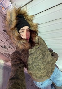 Personne portant une veste camouflage avec une capuche bordée de fourrure et un bonnet noir, debout sur le trottoir près d'un mur couvert de graffitis en plein jour.