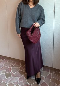 Άτομο που φορά γκρι πουλόβερ με λαιμόκοψη V και σκουρόχρωμη μωβ σατέν φούστα, κρατώντας ένα πλεκτό μπορντό τσαντάκι χειρός, στέκεται σε δάπεδο με πλακάκια με σχέδιο.