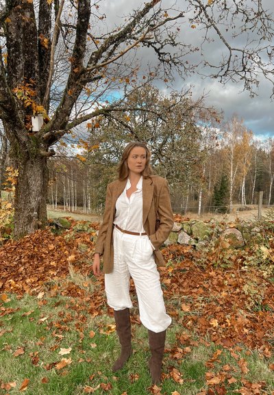 Mujer con conjunto blanco y blazer marrón de pie sobre césped cubierto de hojas otoñales junto a un árbol con ramas desnudas y una caseta para pájaros.