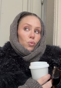 Kobieta w wełnianej chustce na głowie i czarnym futrzanym płaszczu trzymająca kubek z kawą na wynos i okulary przeciwsłoneczne, płatki śniegu na ubraniu, patrząca na bok na zewnątrz.