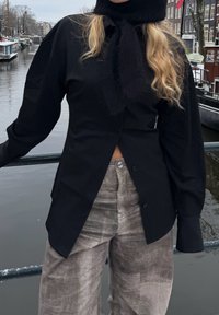 Personne portant une chemise noire sur un pantalon gris, debout près de la rambarde d'un canal avec des bâtiments et des bateaux en arrière-plan par une journée nuageuse.