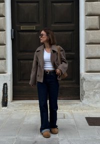 Hnědý dvouřadý kabát, bílý crop top, tmavé rozšířené džíny a béžové nazouvací boty. Opálená kožená kabelka přes rameno dotváří vzhled.