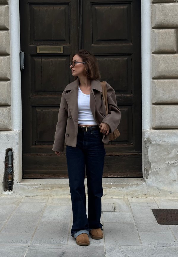 Bruine dubbelgebreefde jas, witte crop top, donkere flared jeans en beige instaplaarzen. Een leren schoudertas in tan kleur maakt de look compleet.