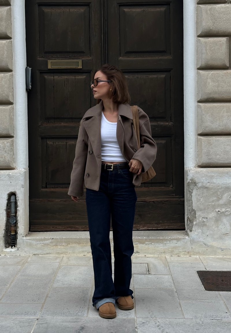 Brown double-breasted coat, white crop top, dark flared jeans, and beige slip-on boots. A tanned leather shoulder bag completes the look.