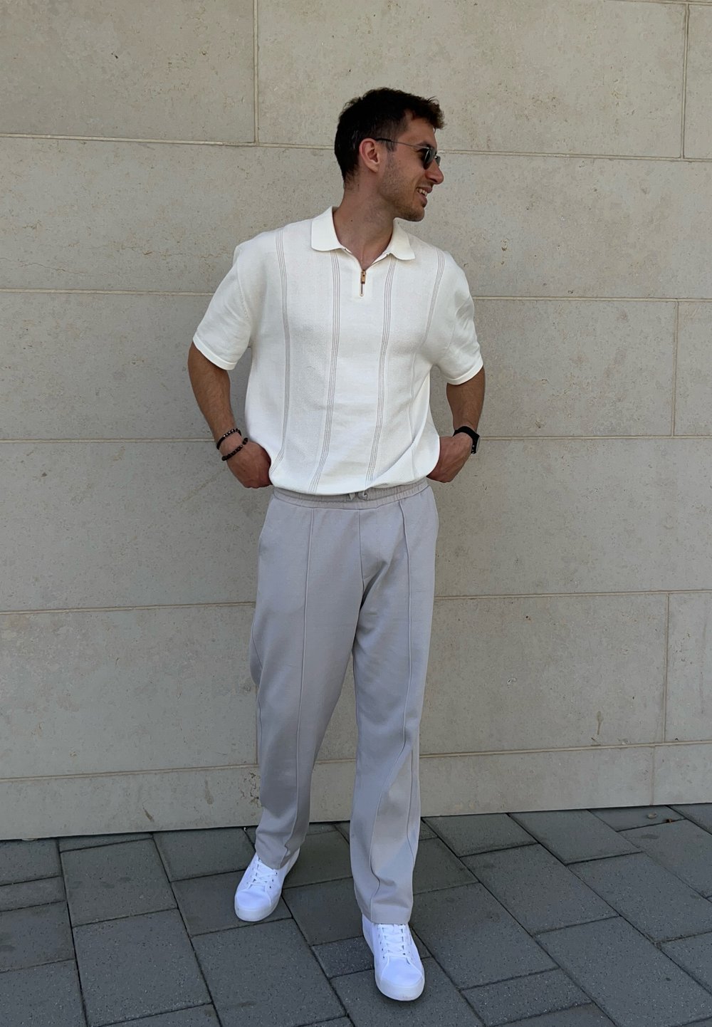 Polo shirt bianco a maniche corte con zip e righe verticali, abbinato a pantaloni grigi chiari a gamba affusolata e sneakers bianche.