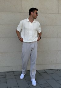 Polo shirt bianco a maniche corte con zip e righe verticali, abbinato a pantaloni grigi chiari a gamba affusolata e sneakers bianche.