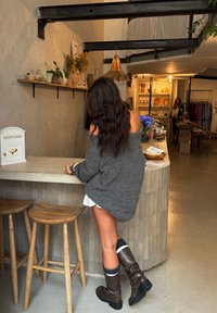 Sivi oversized pleteni pulover, bijela suknja, smeđe čizmice do koljena. Drveni barski stolci i teksturirana kamenita pulta u slabo osvijetljenoj trgovini.