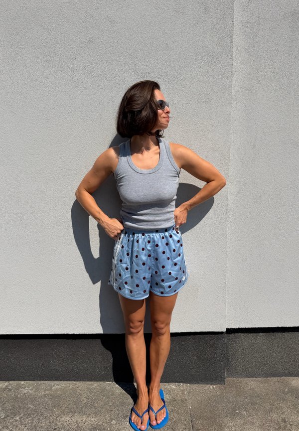 Femme portant des lunettes de soleil, un débardeur gris, un short bleu à pois et des tongs bleues, debout contre un mur gris clair avec les mains sur les hanches.