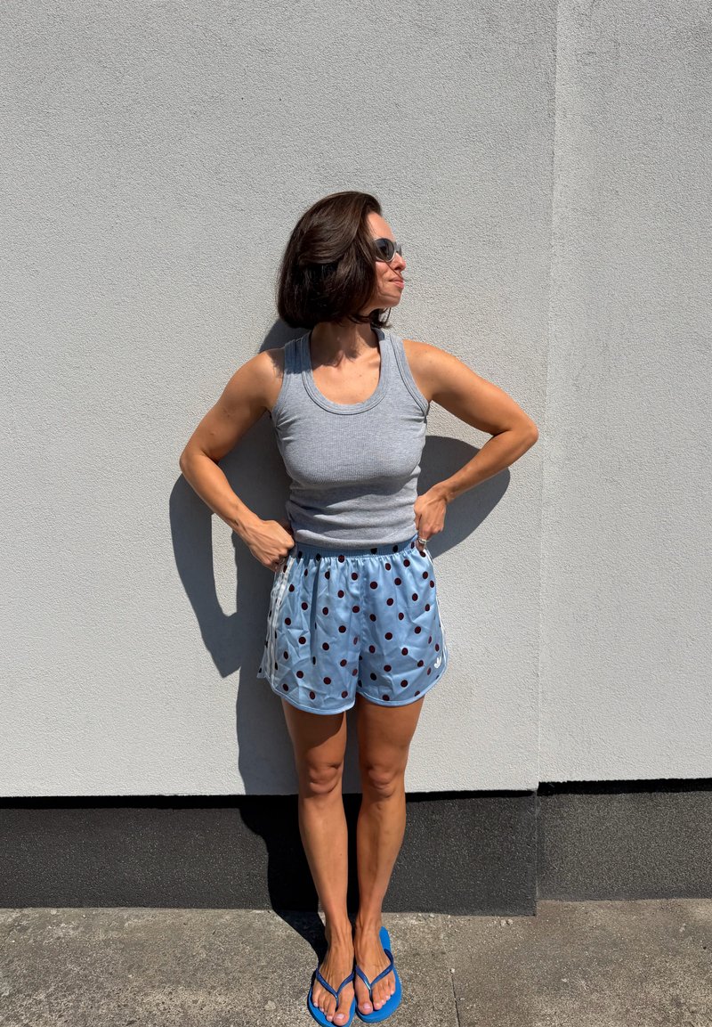 Frau mit Sonnenbrille, grauem Tanktop, blauen Shorts mit Punkten und blauen Flip-Flops steht mit den Händen in der Hüfte vor einer hellgrauen Wand.
