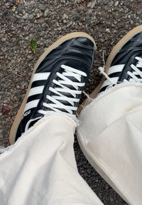 Baskets noires à rayures blanches et lacets, portées avec un pantalon écru effiloché, debout sur un asphalt rugueux avec une petite feuille verte à côté.