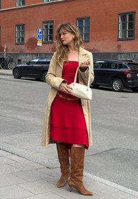 Beige trench coat piros, réteges ruhára, térdig érő barna velúr csizmával, egy kis fehér kézitáskával a kezében, hullámos hajjal és arany ékszerekkel.