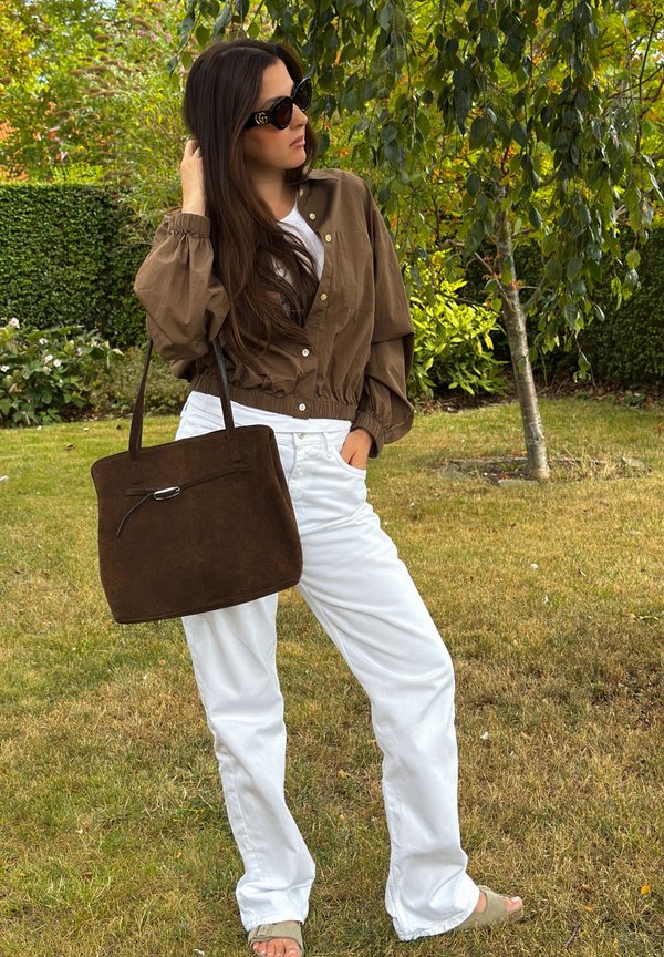 Woman standing outdoors in white pants, brown buttoned jacket, sunglasses, holding a large brown handbag, with trees and grass in the background.