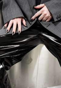 Pantalon en vinyle noir brillant associé à un blazer oversize à carreaux gris. Mains ornées de longs ongles pointus de couleur foncée.