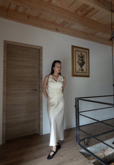 Vestido midi crema con un corpiño ajustado y una falda acampanada, combinado con zapatos planos negros. Pared de paneles de madera y obras de arte enmarcadas de fondo.