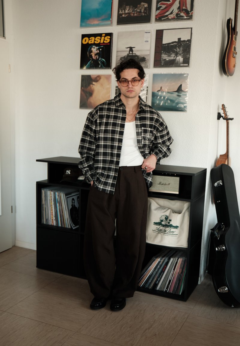Jeune homme portant des lunettes, une chemise à carreaux et un pantalon large, debout à côté de disques vinyles, d'une housse de guitare et de pochettes d'albums encadrées sur un mur blanc.