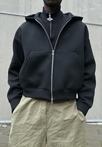 Felpa nera corta con zip frontale, grandi tasche e colletto a coste, abbinata a pantaloni chiari e stropicciati.