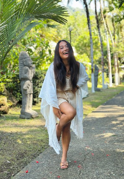 Mujer sonriente con conjunto ligero de punto y chal blanco, doblando una rodilla sobre un camino de jardín rodeado de plantas tropicales y estatuas de piedra.