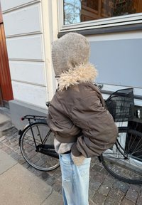 Personne en vêtements chauds avec une capuche en tricot et une veste marron, debout à côté d'un vélo noir devant un bâtiment avec un pavé en pierre.