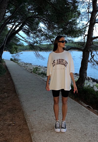 Camiseta blanca de gran tamaño con estampado de "Los Ángeles", combinada con pantalones cortos de bicicleta negros y zapatillas blancas y negras. El fondo incluye árboles y agua.