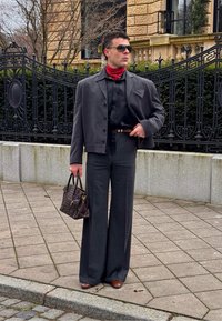 Homme portant un pantalon large à fines rayures, chemise noire, veste courte grise, écharpe rouge, chaussures marron, lunettes de soleil, tenant un sac à main en cuir marron sur un trottoir en ville.